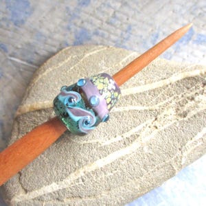 May include: A decorative glass bead with swirling teal and lavender patterns, accented with small blue glass dots. The bead is mounted on a wooden stick, resting on a textured stone surface. The bead features a purple and yellow floral design.