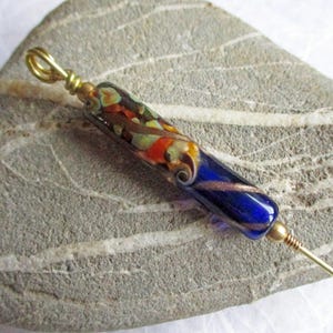 May include: A glass bead with a blue, orange, and brown swirl pattern is attached to a gold-colored pin. The pin is lying on a gray, textured surface.