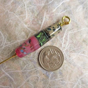 May include: A cylindrical glass bead pendant featuring a multi-colored pattern of pink, green, and dark brown tones, attached to a gold-toned metal wire loop. A United States quarter coin is placed next to the bead for a size reference.