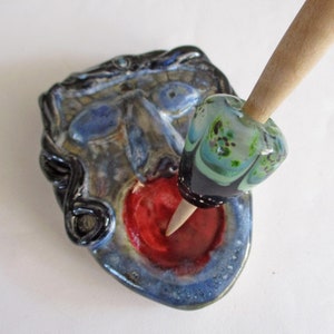 May include: A ceramic face with a red mouth and blue and black glaze. A wooden spindle with a colorful glass bead is resting in the mouth.