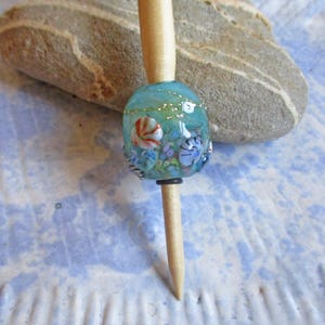 May include: A light-colored wooden hair stick featuring a spherical glass bead. The bead is light blue with embedded multi-colored floral patterns, small white dots, and thin gold-colored lines. The stick has a tapered, pointed end.