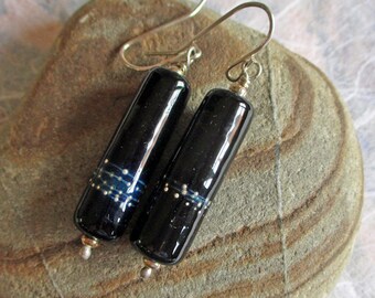 May include: A pair of black glass earrings with silver-colored hooks. Each earring is a cylindrical shape with a band of blue and white dots. The earrings are resting on a gray stone.