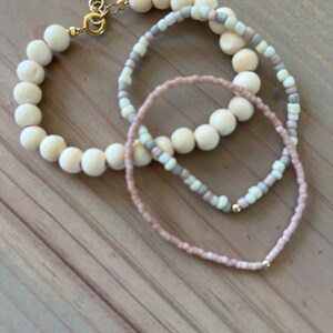 May include: Three bracelets made of wooden beads and small pink, white, and grey beads. The bracelets are layered on top of each other.