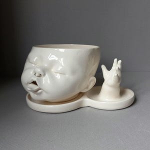 May include: A glossy off-white ceramic planter shaped like a baby head with closed eyes, a small silver nose ring, and an open mouth with a protruding tongue. Attached to the side is a small ceramic hand reaching upward from the base tray.