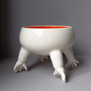 May include: A white ceramic bowl with a smooth, glossy finish and a vibrant orange interior. The bowl is supported by four sculpted legs that terminate in detailed hand-shaped feet, giving the vessel a distinct, elevated, and stable appearance.
