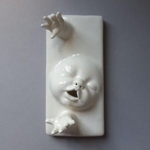 May include: A white ceramic wall sculpture featuring a stylized face with closed eyes and an open mouth, with two hands reaching out. The sculpture is rectangular and has a glossy finish.