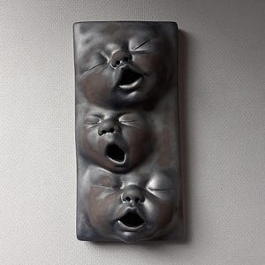 May include: A rectangular, dark gray ceramic wall hanging featuring three stylized baby faces. Each face has closed eyes and an open mouth, creating a textured, artistic effect. The piece is mounted on a light gray surface.