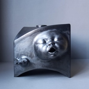 May include: A metallic gray sculpture of a baby's face emerging from a rectangular form. The baby's eyes are closed, and its mouth is open. A small hand extends from the side. The sculpture has a small, circular opening on top.