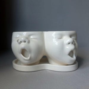 May include: A white ceramic double planter shaped like two yawning faces, with one face featuring a small silver-colored hoop ring in the nose. The planter sits on a matching white ceramic tray that connects both head-shaped vessels.