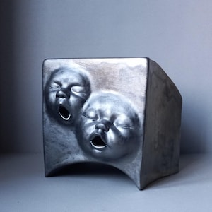 May include: A silver-colored, cuboid sculpture featuring two embossed baby faces with open mouths and closed eyes. The artwork has a metallic sheen and a curved base, creating a unique and artistic piece.