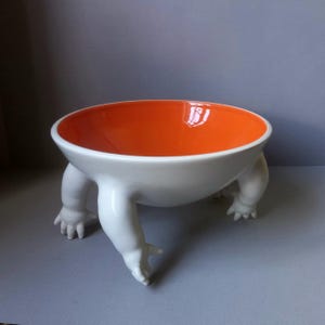 May include: A white ceramic bowl with an orange interior, supported by three stylized legs that resemble human limbs. The bowl has a glossy finish and is set against a neutral background. This unique piece is a decorative item.
