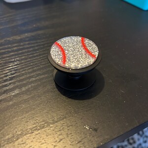 May include: A black phone grip with a baseball design. The round top features a silver glitter background with red stitching details. The grip is designed to attach to the back of a phone for easier handling.