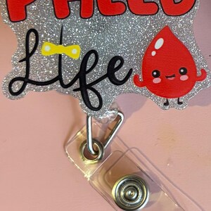 Puede incluir: Un carrete de insignia brillante con las palabras "PHLEB Life" en rojo y negro. Una linda gota de sangre de dibujos animados está a la derecha. El carrete de la insignia tiene una correa de plástico transparente y un clip de metal.