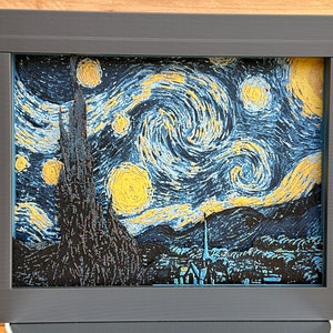 May include: A framed print of Vincent van Gogh's "Starry Night" painting. The painting features a swirling night sky with a bright yellow moon and stars, and a dark silhouette of a village in the foreground.