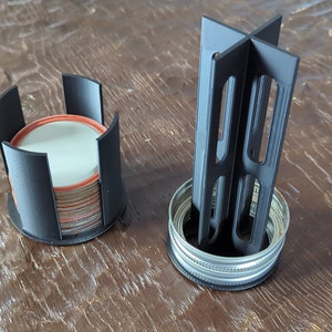 May include: Two black plastic organisers for storing jar lids. One organiser is a round cylinder with slots for stacking lids. The other organiser is a cross-shaped stand with slots for holding lids upright.