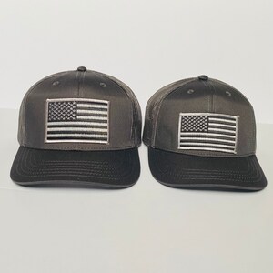 May include: Two black and gray trucker hats with a white embroidered American flag patch on the front of each hat.