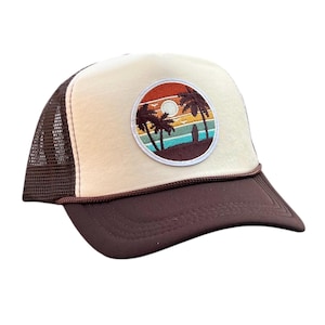 May include: A trucker hat with a brown brim and mesh back. The front panel is beige with a circular patch featuring a sunset scene with palm trees, a surfboard, and a sun. The hat has a brown rope detail along the brim.