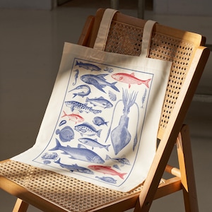 May include: A cream-colored canvas tote bag featuring a rectangular print of various marine life illustrations. The design includes blue and red fish, a large central squid, a crab, and shells arranged within a thin blue border on the fabric.