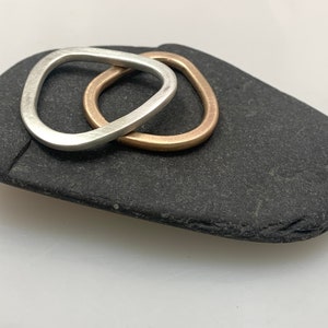 Puede incluir: Dos anillos abstractos, uno plateado y otro dorado, entrelazados sobre una piedra gris oscura. Los anillos tienen una forma suave y orgánica. La piedra tiene una textura rugosa y está sobre un fondo blanco. La joyería es minimalista y moderna.