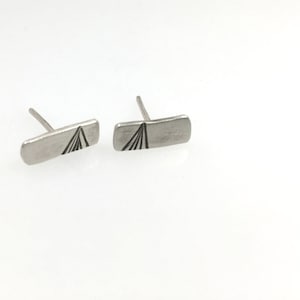 May include: A pair of silver stud earrings with a geometric design. Each earring features a rectangular shape with a black line design that resembles a mountain peak.