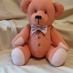 May include: A handmade peach-colored teddy bear with a bow tie and button-down shirt. The bear's paws and inner ears feature a white and orange checkered pattern. The bear is sitting on a cream-colored surface.