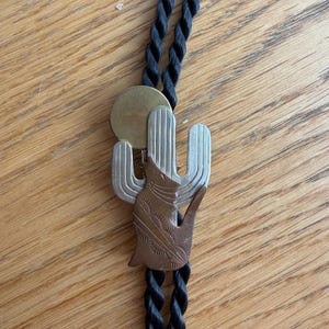 May include: A bolo tie featuring a black braided cord and a metal slide. The slide depicts a silver-toned saguaro cactus, a brass-toned circular moon, and a copper-toned howling coyote silhouette with etched decorative line details.