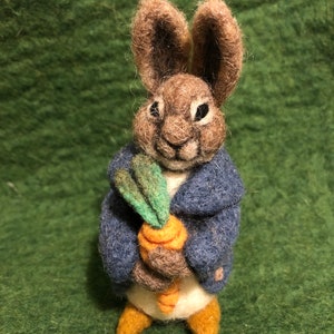 May include: A handcrafted felt bunny figurine, wearing a blue jacket and holding a carrot. The bunny has brown fur, white belly, and tan feet. The carrot has an orange body and green leaves. The background is a solid green.