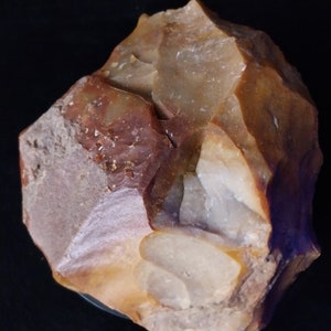 May include: A large, irregularly shaped, brown, tan, and white stone tool with a rough, chipped surface. The stone has a purple hue on the bottom edge.