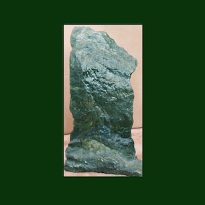 May include: A large, dark green, rough-textured rock with a jagged edge. The rock is standing upright on a light brown surface.