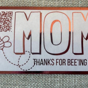 May include: A rectangular card with a rose gold border and the word "MOM" in large, rose gold letters. Below, the phrase "THANKS FOR BEE'ING YOU!" is written. A bee graphic and QR code are also present.