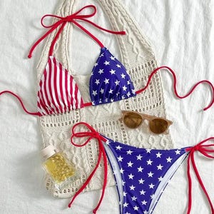 May include: A patriotic bikini set with a red, white, and blue color scheme. The top features red and white stripes on one cup and blue with white stars on the other. The bottoms are blue with white stars. Red ties accent the bikini. A cream-colored tote bag, sunglasses, and a small bottle are also in the image.