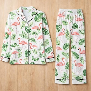 May include: White pyjama set featuring a flamingo and tropical leaf design. The long-sleeved top has black trim and pockets. The matching trousers have an elasticated waist. The set is made of a soft, lightweight fabric.