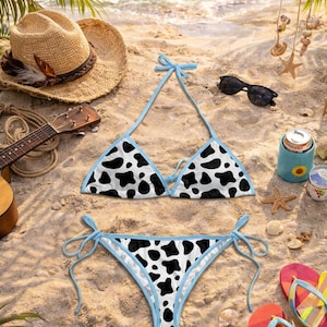 May include: A cow-print bikini set with a light blue trim, lying on a sandy beach. The bikini top and bottom feature a black and white pattern. Other beach accessories include a straw hat, sunglasses, and flip-flops.