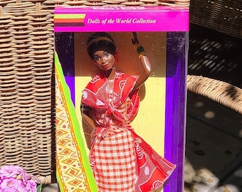 Puede incluir: Una muñeca Barbie keniana en caja de la colección Dolls of the World. La muñeca lleva un atuendo con estampado rojo y blanco con un ribete dorado y verde. La caja es morada y amarilla, y la muñeca se exhibe en una silla de mimbre junto a una maceta morada.