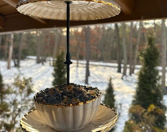May include: A white porcelain bird feeder with gold trim, suspended from a black metal rod. The feeder is shaped like a teacup and saucer, filled with birdseed. A decorative bird figurine sits atop the feeder's canopy, and a crystal hangs below.