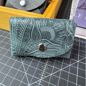 May include: A teal leather wallet with an embossed floral design and a silver snap closure.