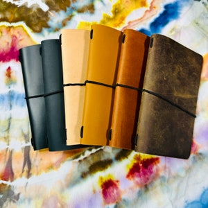 May include: Five leather-bound journals in different shades of brown, black, and tan, each with a black elastic band closure.