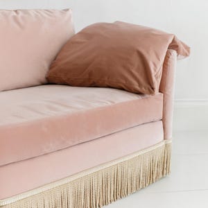 May include: Close-up of a pink velvet sofa with a matching pillow and a brown pillow. The sofa has a fringe trim along the bottom edge. The sofa is set against a white background.