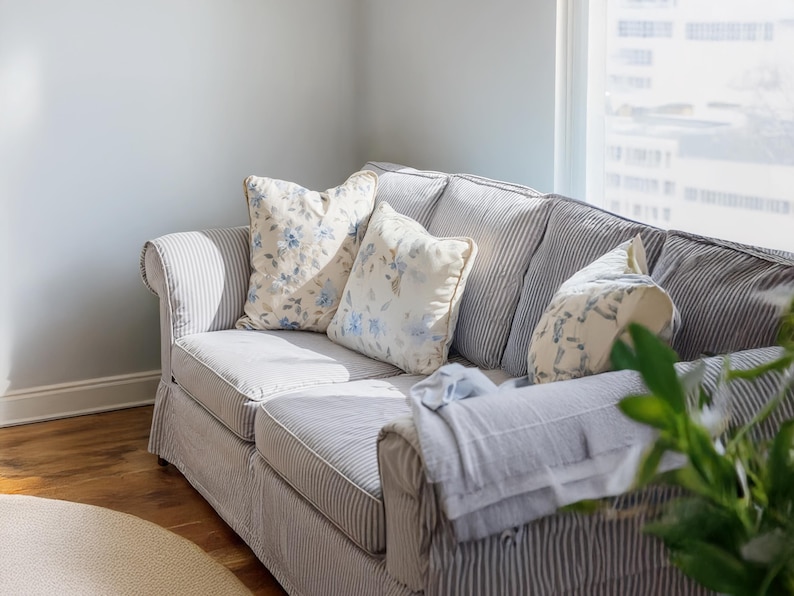 Pu&ograve; includere: Un divano colorato chiaro con un motivo a righe e cuscini floreali. Il divano ha una base a gonna e braccioli arrotolati. Una coperta azzurra &egrave; drappeggiata sul braccio. La stanza ha una finestra e pavimenti in legno.