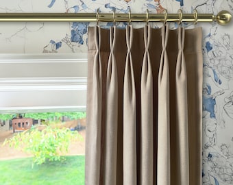 May include: Beige curtains with pleats hanging from a gold curtain rod. The curtains are in front of a window with a view of a green lawn and trees.