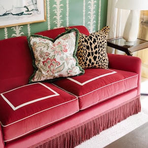 Könnte beinhalten: Ein rotes Samtsofa mit zwei dekorativen Kissen. Ein Kissen hat ein Blumenmuster mit grüner Bordüre, das andere einen Leopardenmuster. Das Sofa hat eine weiße Bordüre und einen Fransenbesatz am unteren Rand.
