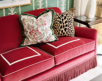 May include: A red velvet sofa with two decorative pillows. One pillow features a floral pattern with green trim, and the other has a leopard print. The sofa has white trim and a fringe along the bottom.