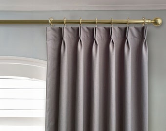 May include: Gray pleated curtains hanging from a gold curtain rod with finials. The curtains are drawn and the rod is mounted above a window with white blinds.