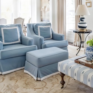 May include: Two pale blue armchairs and an ottoman with white trim. The chairs have square cushions with white borders. A round side table with a lamp and a patterned bench are also in the room. The room has a neutral colour palette.