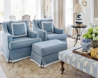 May include: Two light blue armchairs and a matching ottoman with white trim. The chairs have square cushions with white borders. A patterned bench and a round side table with a lamp are also visible in the room.