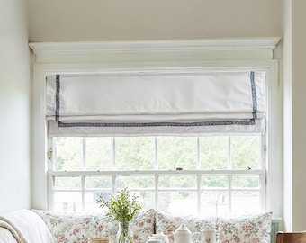 May include: White Roman shade with a navy blue patterned border, installed above a multi-pane window. The window is in a room with a floral patterned sofa and a vase of greenery.