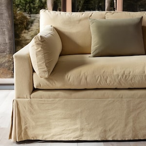 Puede incluir: Un sofá tapizado beige con dos cojines a juego y un cojín verde oliva. El sofá tiene una base con falda y está colocado frente a una ventana. El diseño es simple y moderno, adecuado para una sala de estar.