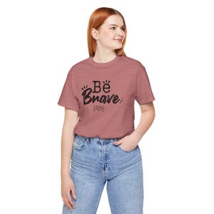 Op de afbeelding: Een oudroze t-shirt met de tekst "Be Brave" in zwart schrift. Het shirt heeft een ronde hals en korte mouwen. De tekst wordt vergezeld door pootafdrukken en een hartsymbool.