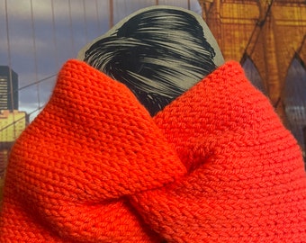 May include: Bright orange knitted headband with a twisted knot design. The headband is made of a thick, textured yarn. The background features a print of the Brooklyn Bridge.
