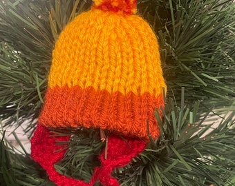 May include: A miniature, hand-knitted hat ornament in shades of yellow, orange, and red. The hat features ear flaps and a pom-pom on top. The ornament is displayed on a green Christmas tree.